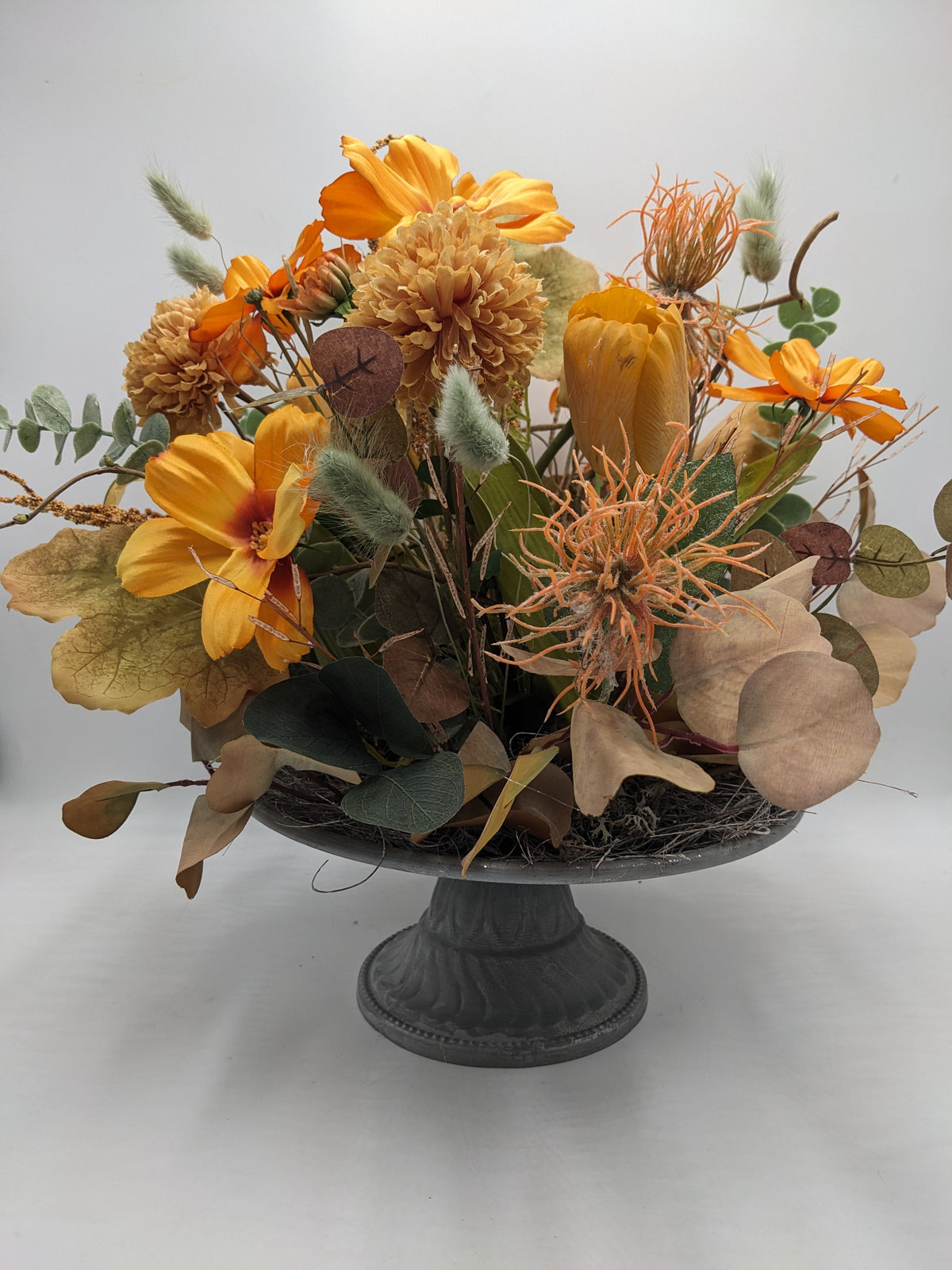 Orangefarbenes Kunstblumengesteck mit gelber Tulpe, Gräsern und getöntem Laub auf grauer Fußschale, seitlich