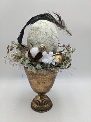 Ostergesteck im goldenen Pokal mit hellgrünem Ei, goldener Schnecke, Baumwolle und dunklen Federn