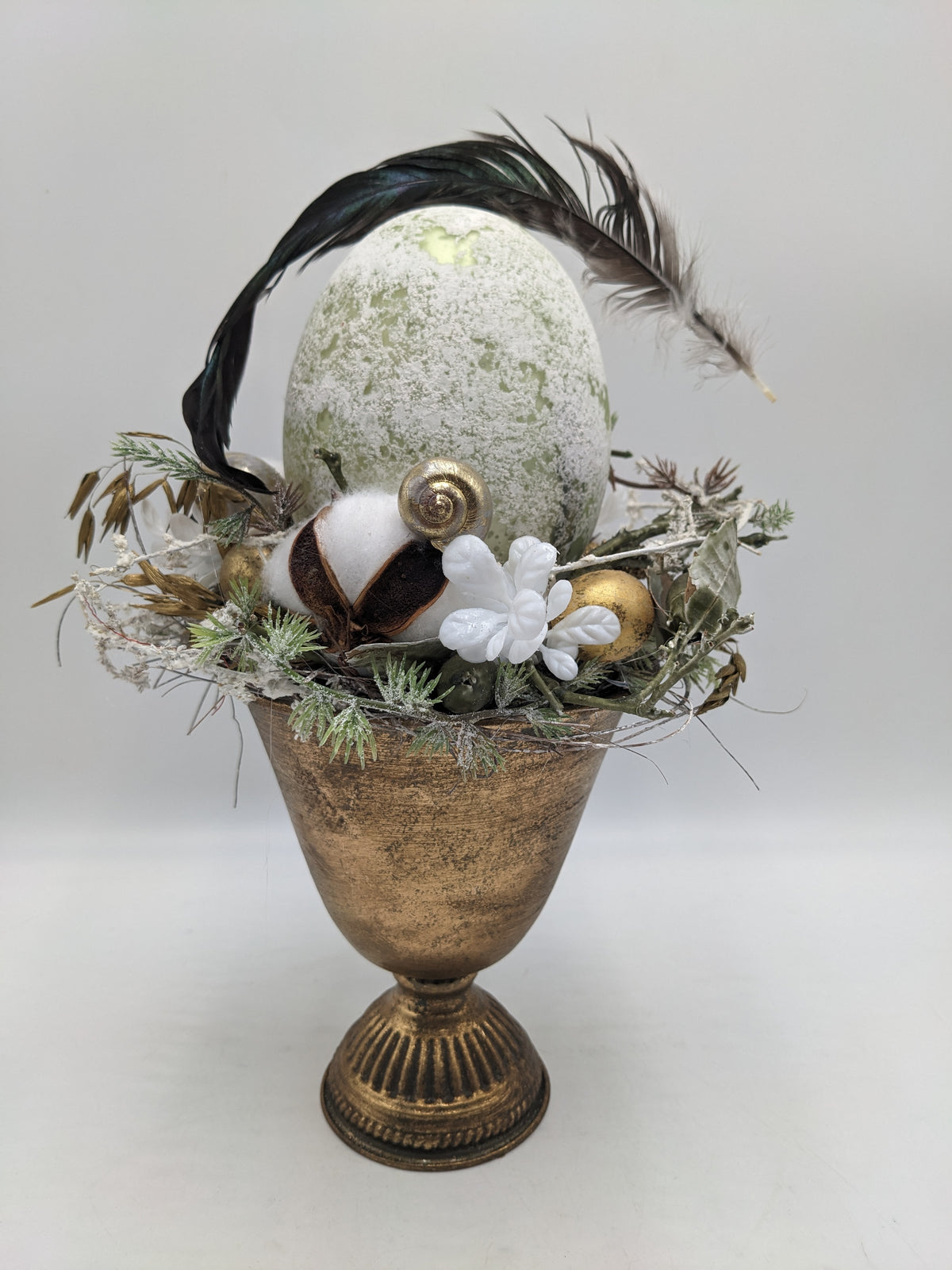 Ostergesteck im goldenen Pokal mit hellgrünem Ei, goldener Schnecke, Baumwolle und dunklen Federn