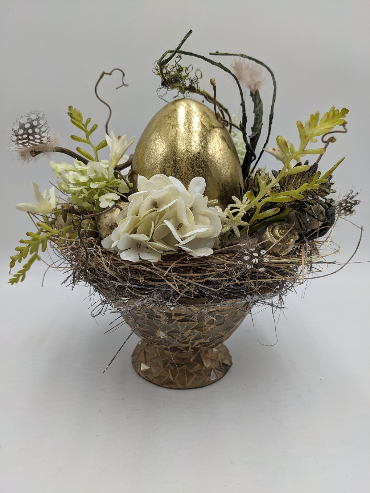 Ostergesteck mit großem goldenem Ei, cremeweißer Hortensie, Hyazinthen und Federn in Mosaik-Fußschale