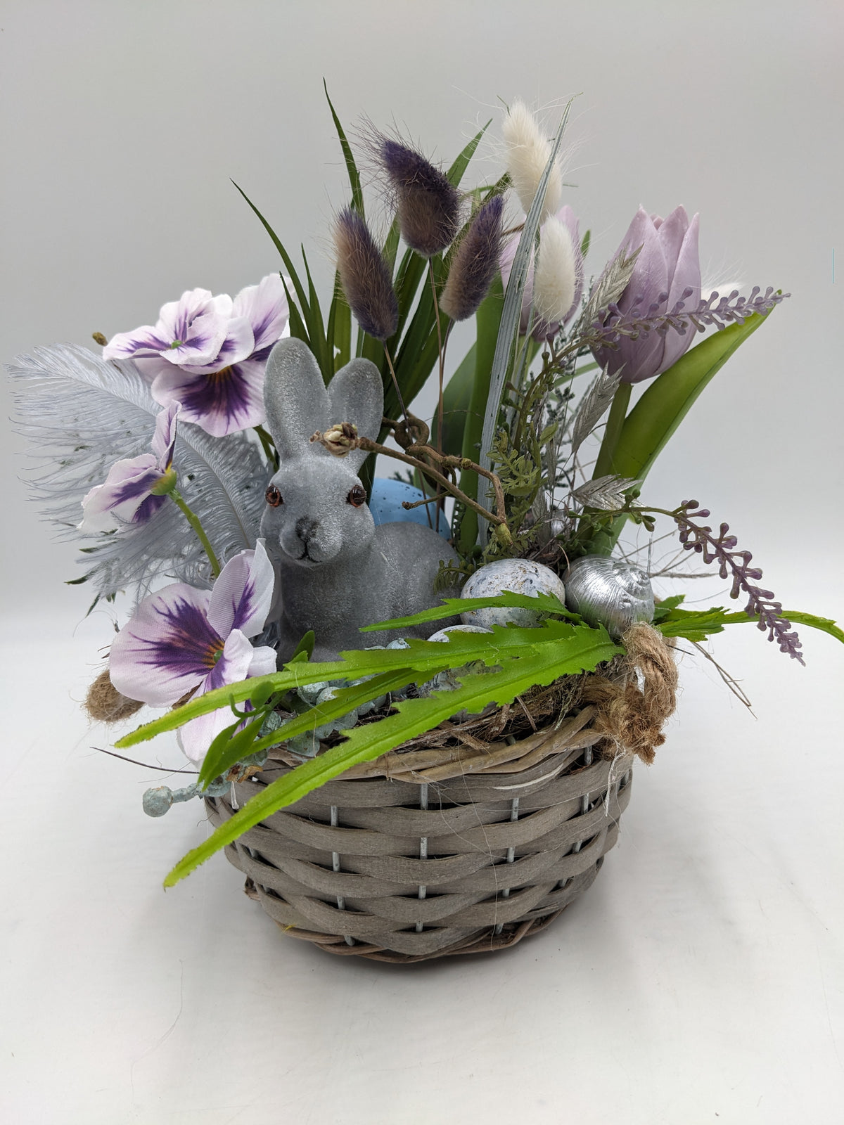 Ostergesteck im grauen Weidenkorb mit grauem Hasen, lila Tulpen, weiß-violetten Stiefmütterchen und Federn