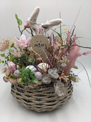 Ostergesteck im Weidenkorb mit Holzhase, rosa Magnolien, Federn und bunten Eiern, von vorn fotografiert