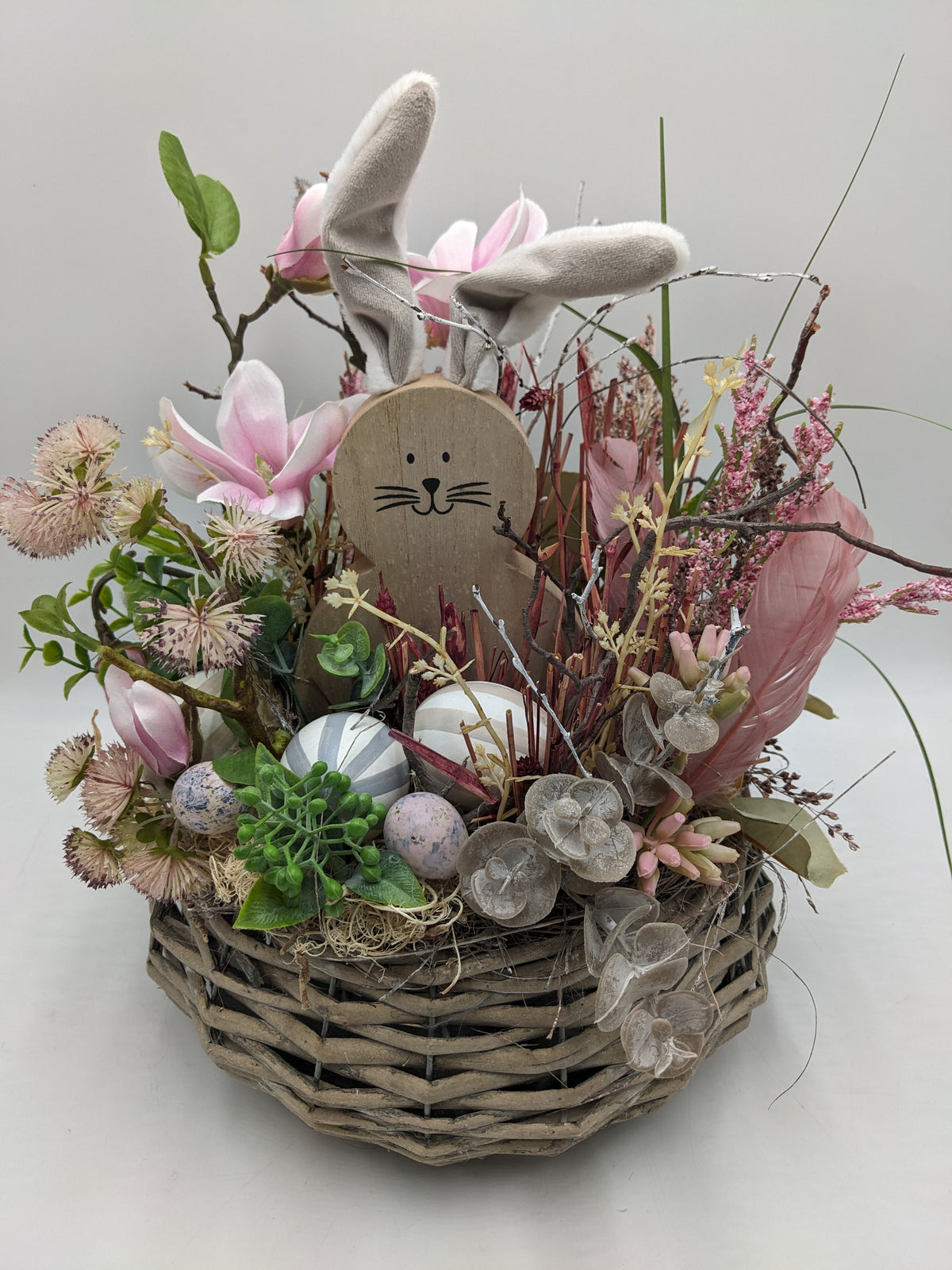 Osterkorb mit Holzhase, Stoffohren, rosa Heidekraut, Silberblatt und Ostereiern zwischen Eukalyptus und Heu