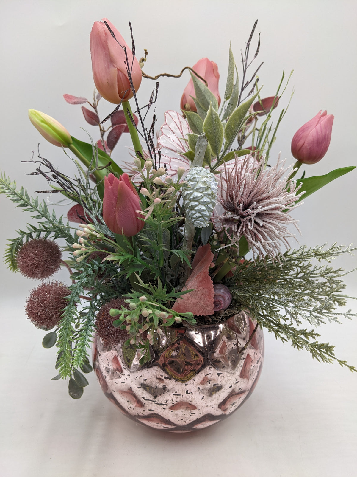 Frühlingsgesteck mit rosa Tulpen, Schmetterling, Zapfen und Gräsern in bauchigem Glastopf in Rosé
