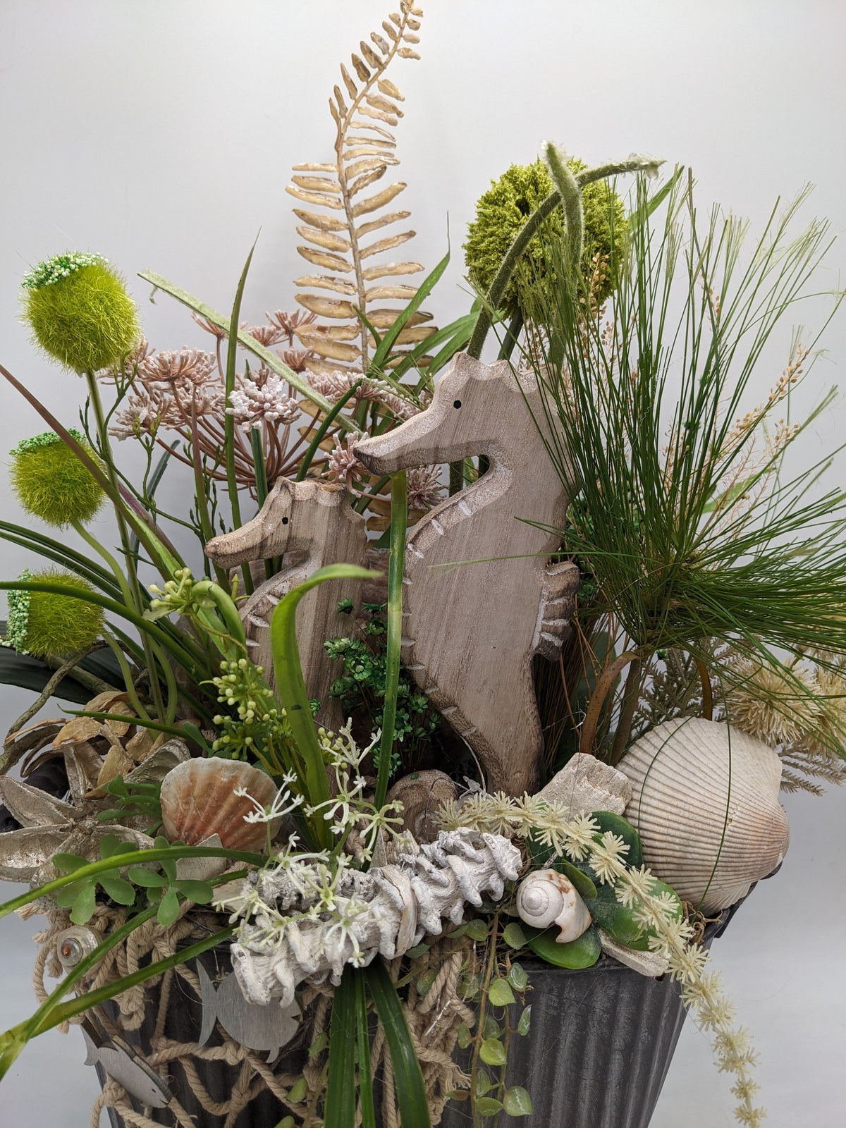 Nahaufnahme: zwei Seepferdchen aus Holz zwischen Gräsern, Farn, Muscheln, Schnecke und weiß gekalktem Korallenzweig