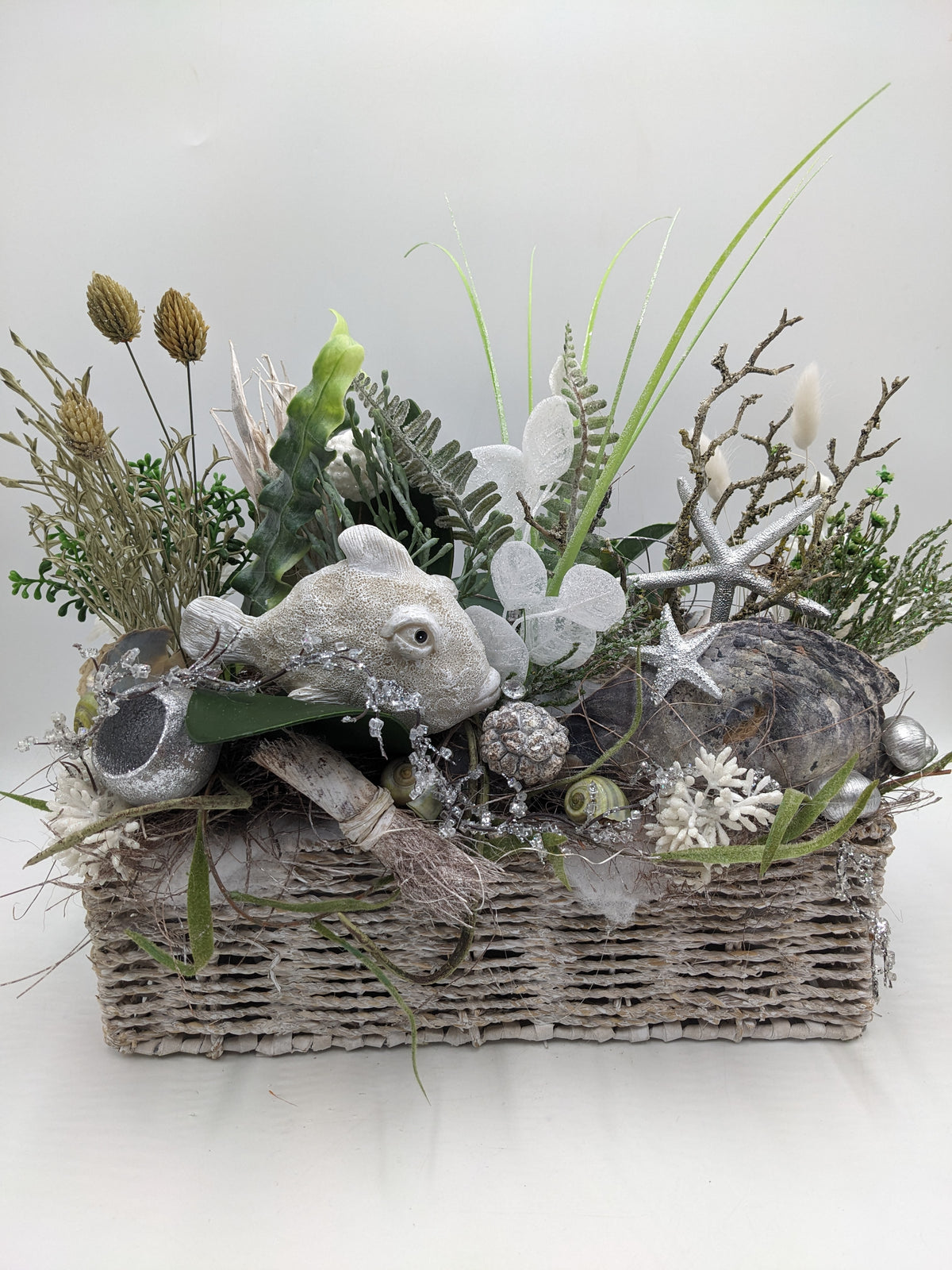 Maritimes Sommergesteck im Korb mit weißem Fisch, silbernen Seesternen, Muscheln, Seetang und Gräsern