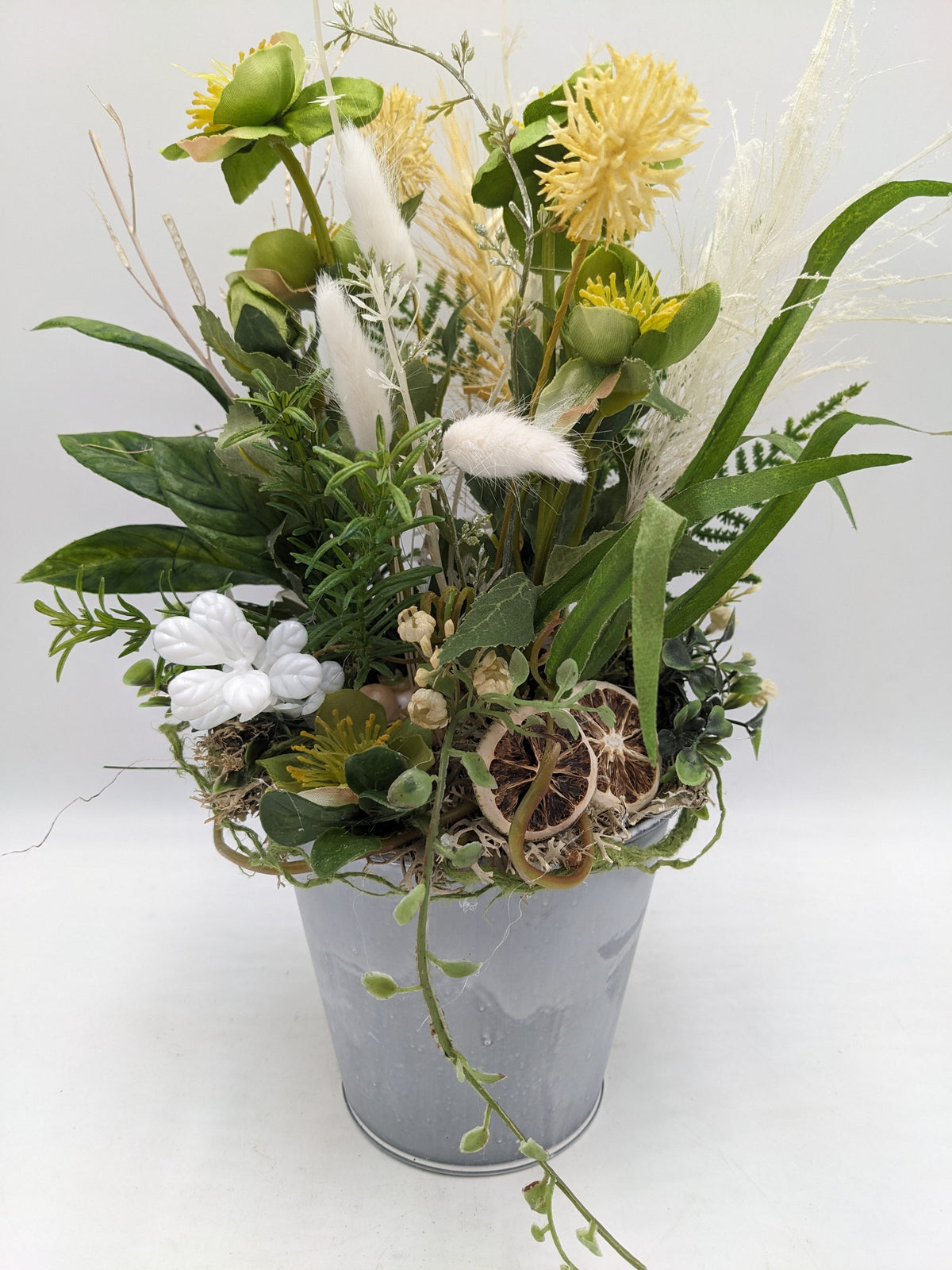 Sommergesteck im Zinktopf mit gelben Blüten, grünen Lenzrosen, weißen Gräsern und getrockneten Zitronenscheiben