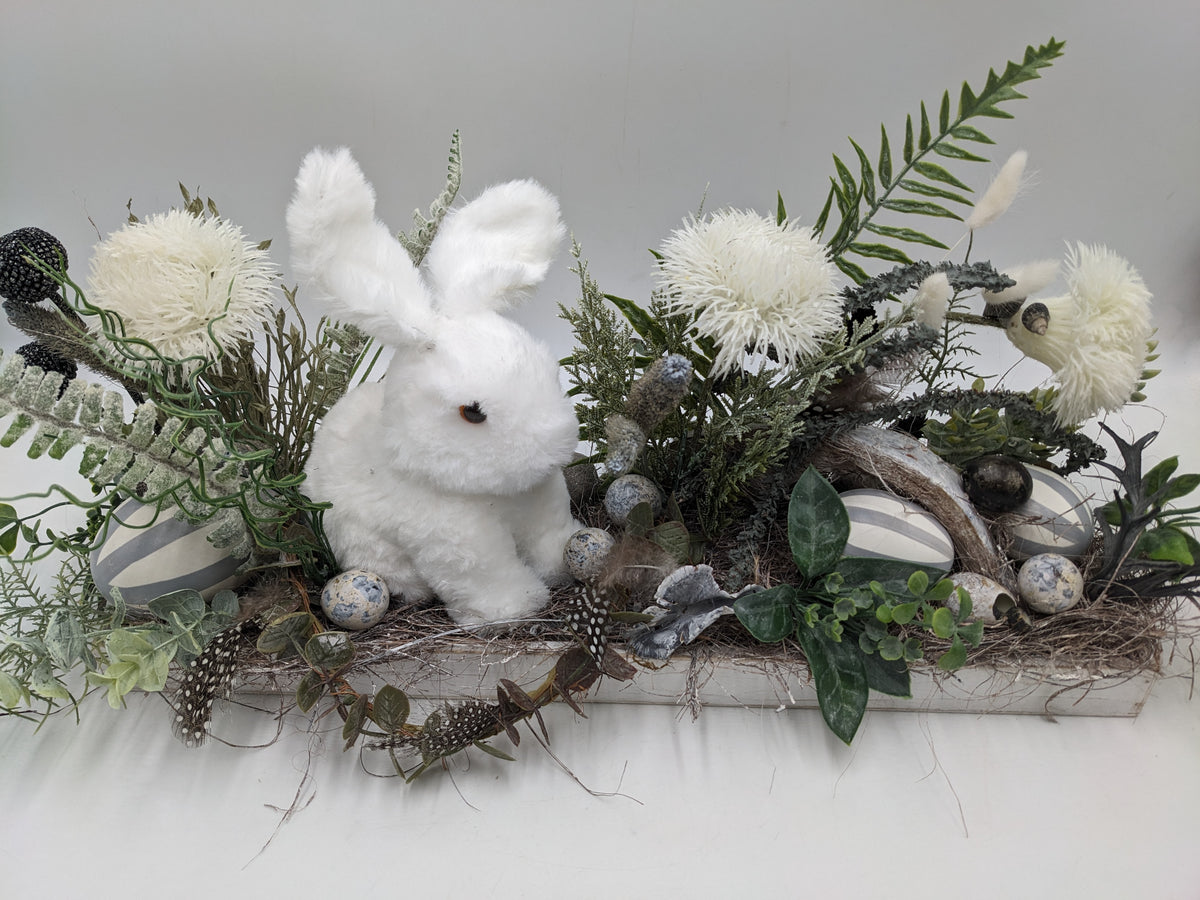 Ostergesteck von vorn: weißer Hase zwischen cremeweißen Blüten, Federn, Nest und gesprenkelten Eiern