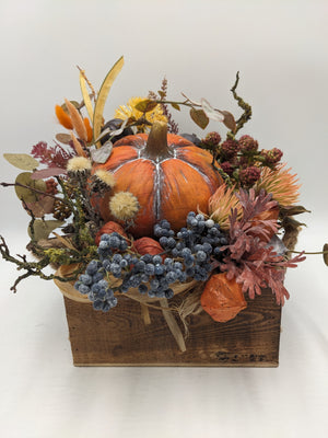 Herbstgesteck in Holzlade mit orangefarbenem Kürbis, blauen Beeren, Physalis, gelber Chrysantheme und Zweigen