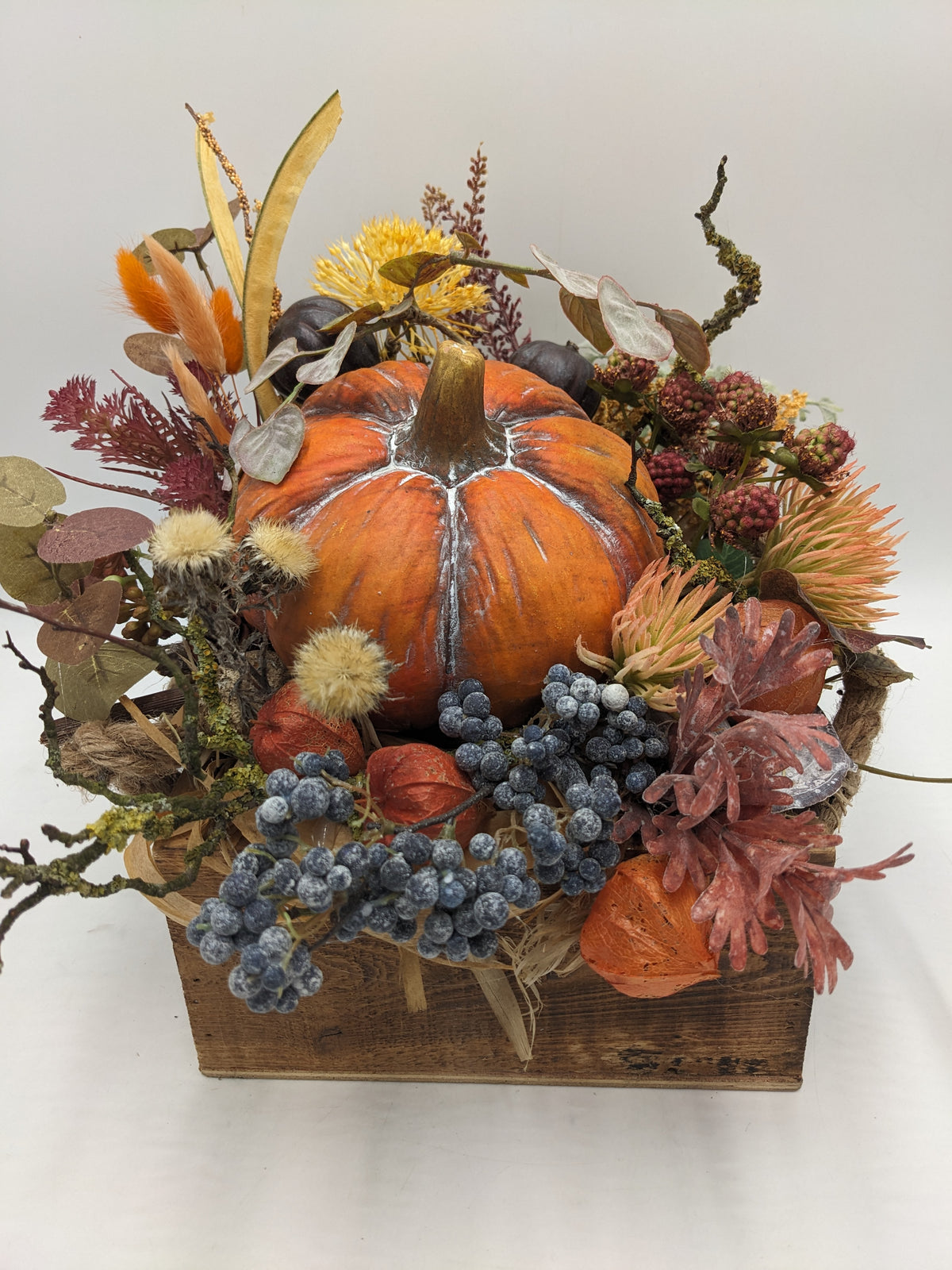 Herbstgesteck von vorn: großer Kürbis in Holzkiste, blau bereifte Beerentrauben, rotes Laub und helle Disteln