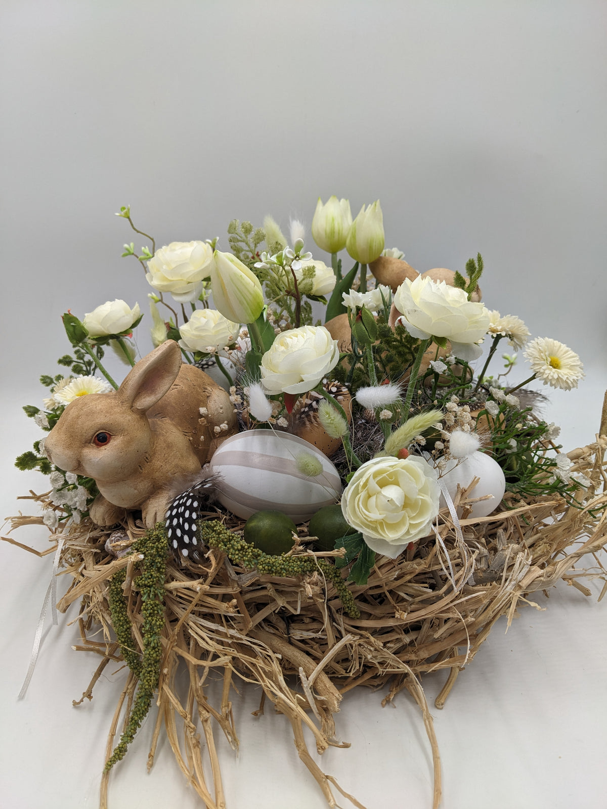 Osterkranz aus Stroh mit braunem Keramikhasen, cremeweißen Ranunkeln, Tulpen, Bellis, Eiern und Federn