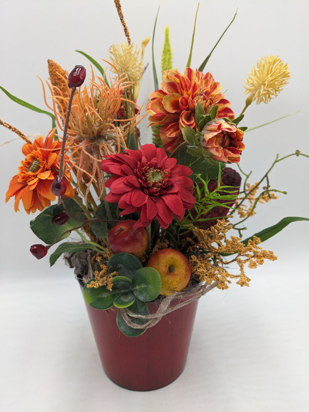 Herbstgesteck von vorn: rote und orange Gerbera, gelbe Rispen, zwei Äpfel und grüne Sukkulente am Topfrand