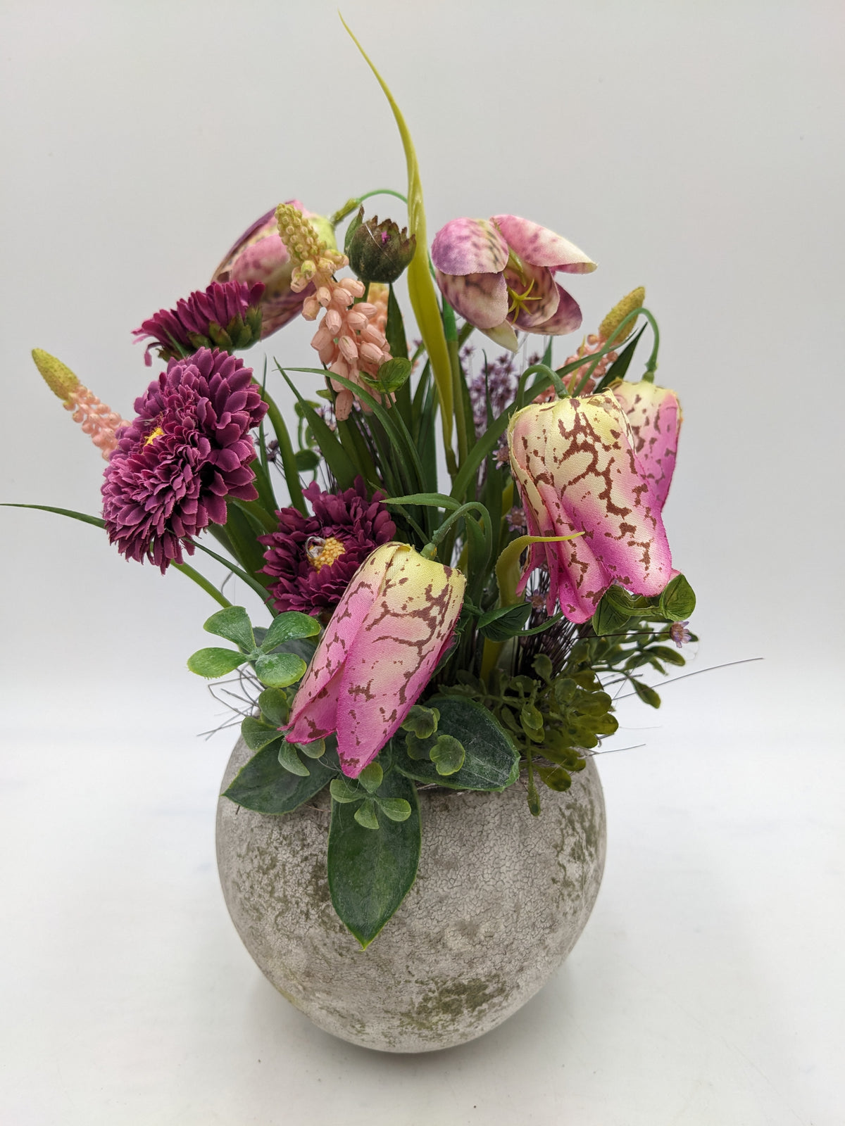 Gesteck mit rosa Schachbrettblumen, lila Bellis und Gräsern in rundem Topf in Steinoptik