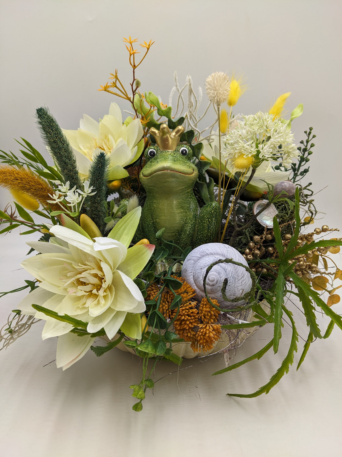 Sommergesteck in heller Schale mit grünem Froschkönig, weißer Schnecke, cremeweißen Seerosen und Gräsern