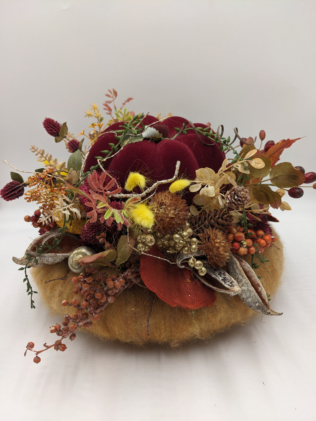 Herbstgesteck mit bordeauxrotem Samtkürbis auf ockerfarbenem Kürbis, dazu Beeren, Zapfen und gelbe Gräser