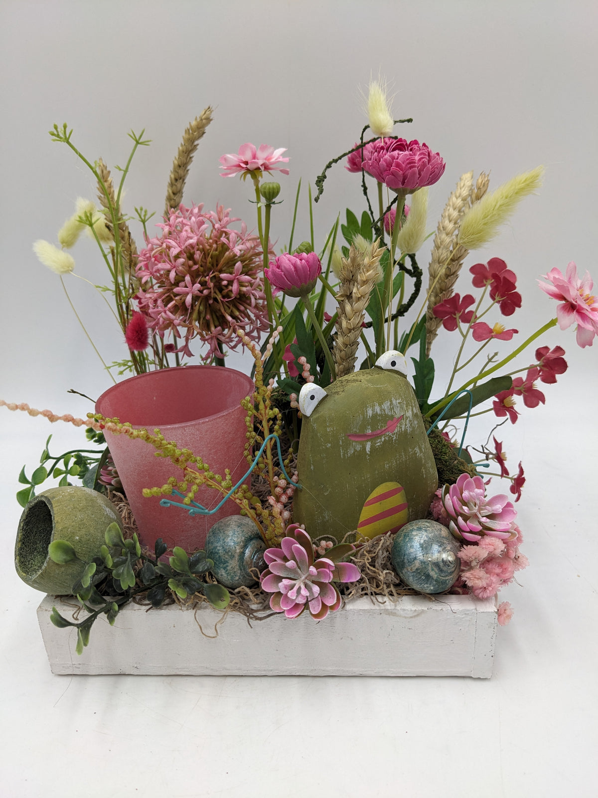 Tischgesteck im weißen Holztablett mit rosa Teelichtglas, grüner Froschfigur, rosa Blüten, Ähren und Gräsern
