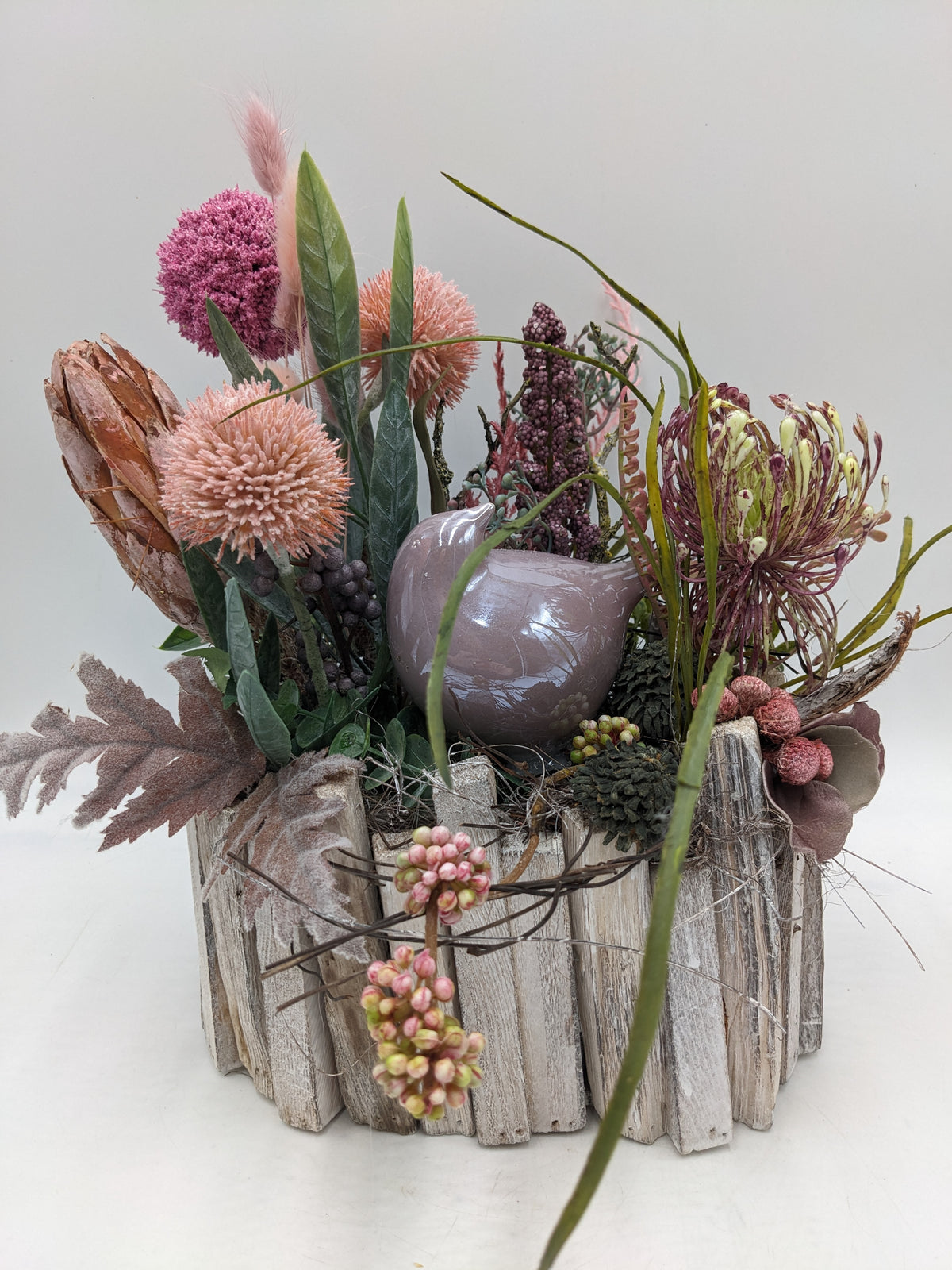 Sommergesteck im Holztopf mit rosa Kugelblüten, Protea, Spider-Blüte und altrosa Keramikvogel