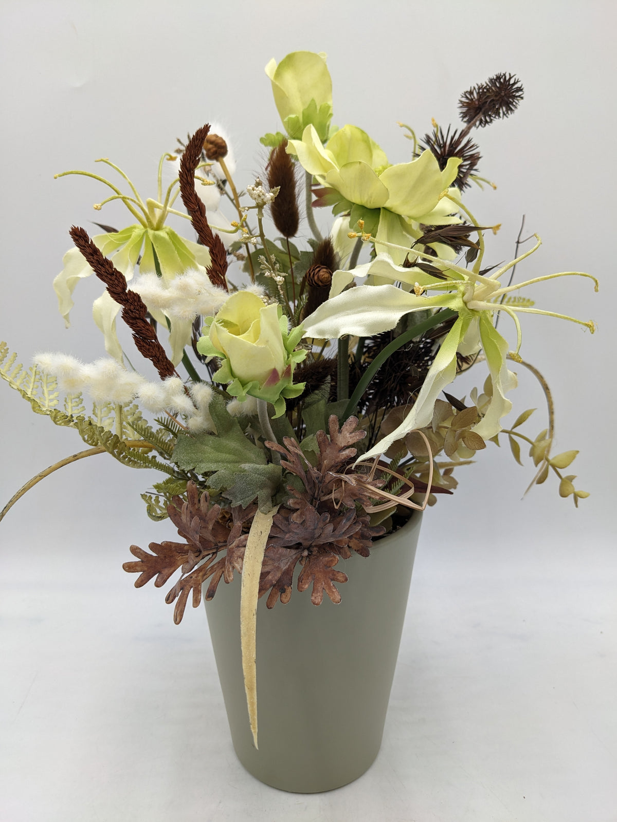 Sommergesteck mit hellgrünen Anemonen, cremefarbener Gloriosa, braunen Disteln und Gräsern in graugrünem Topf
