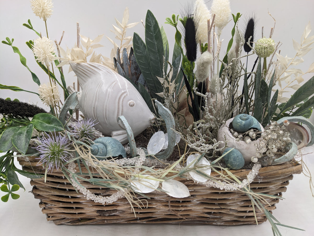 Nahaufnahme des Gestecks: weißer Fisch, Muscheln, Perlenzweige und violette Distel über dem Korbrand