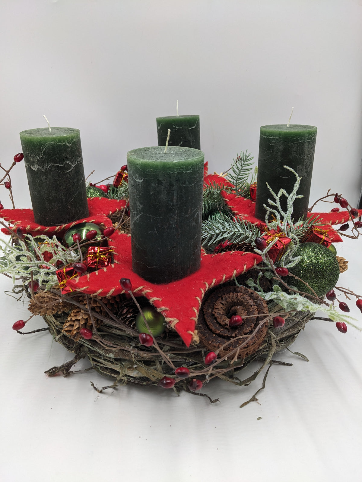 Adventskranz von der Seite: vier dunkelgrüne Stumpenkerzen auf roten Filzsternen, grüne Kugeln, Zapfen und rote Beeren