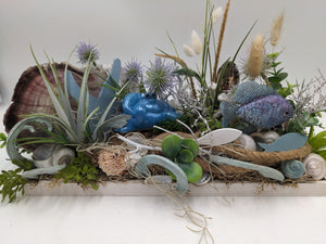 Maritimes Sommergesteck im weißen Holztablett mit Muschel, blauer Krabbe, Fisch, Schnecken und Gräsern