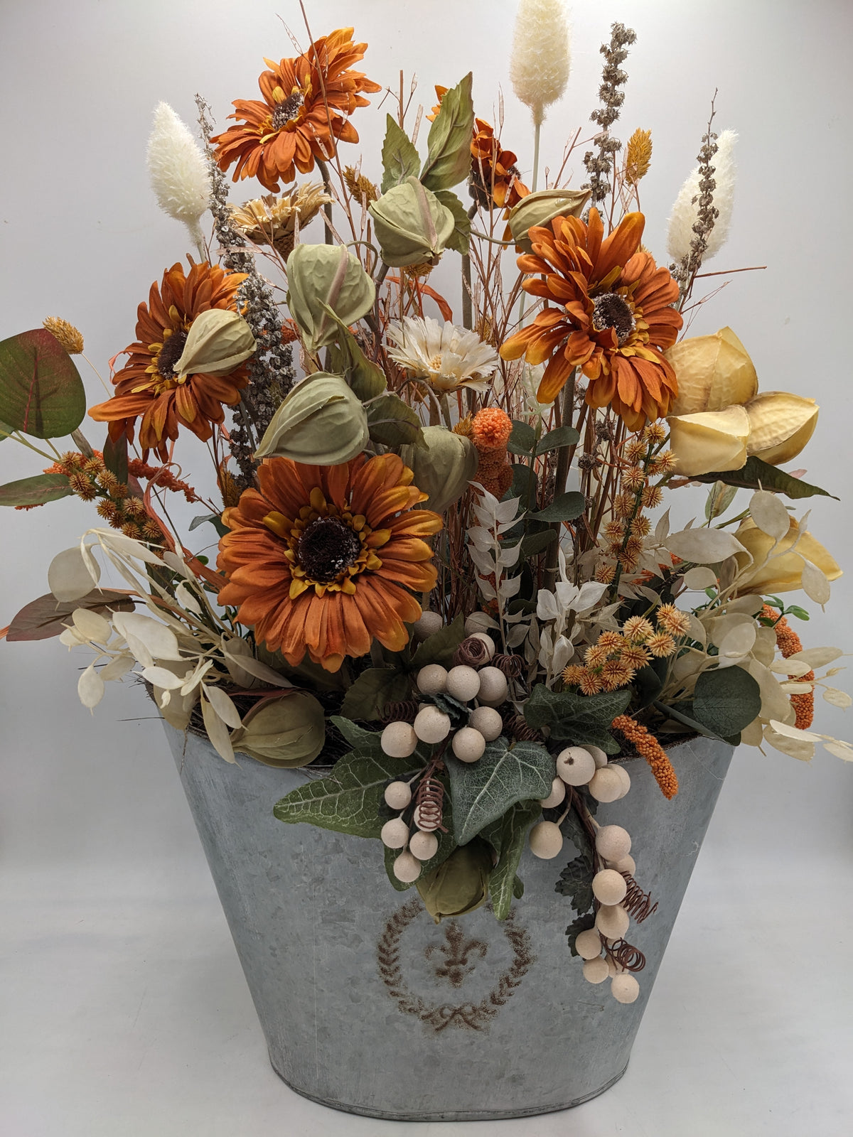 Großes Gesteck mit orangefarbenen Gerbera, grünen Physalislaternen und hellen Gräsern in grauem Zinktopf
