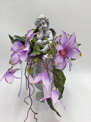 Trauergesteck mit grauer Engelfigur, lila Clematisblüten und Ranken in silberfarbenem Pokal mit Satinband