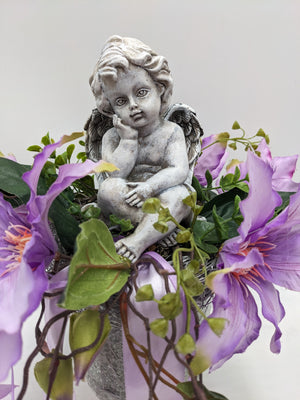 Nahaufnahme der sitzenden Engelfigur zwischen lila Clematisblüten, grünen Blättern und hellem Satinband