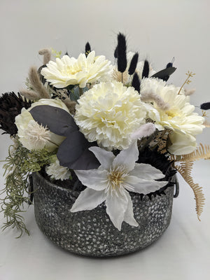 Sommergesteck mit weißen Nelken, Gerbera und Clematis, schwarzen Gräsern und Zapfen in grauer Keramikschale