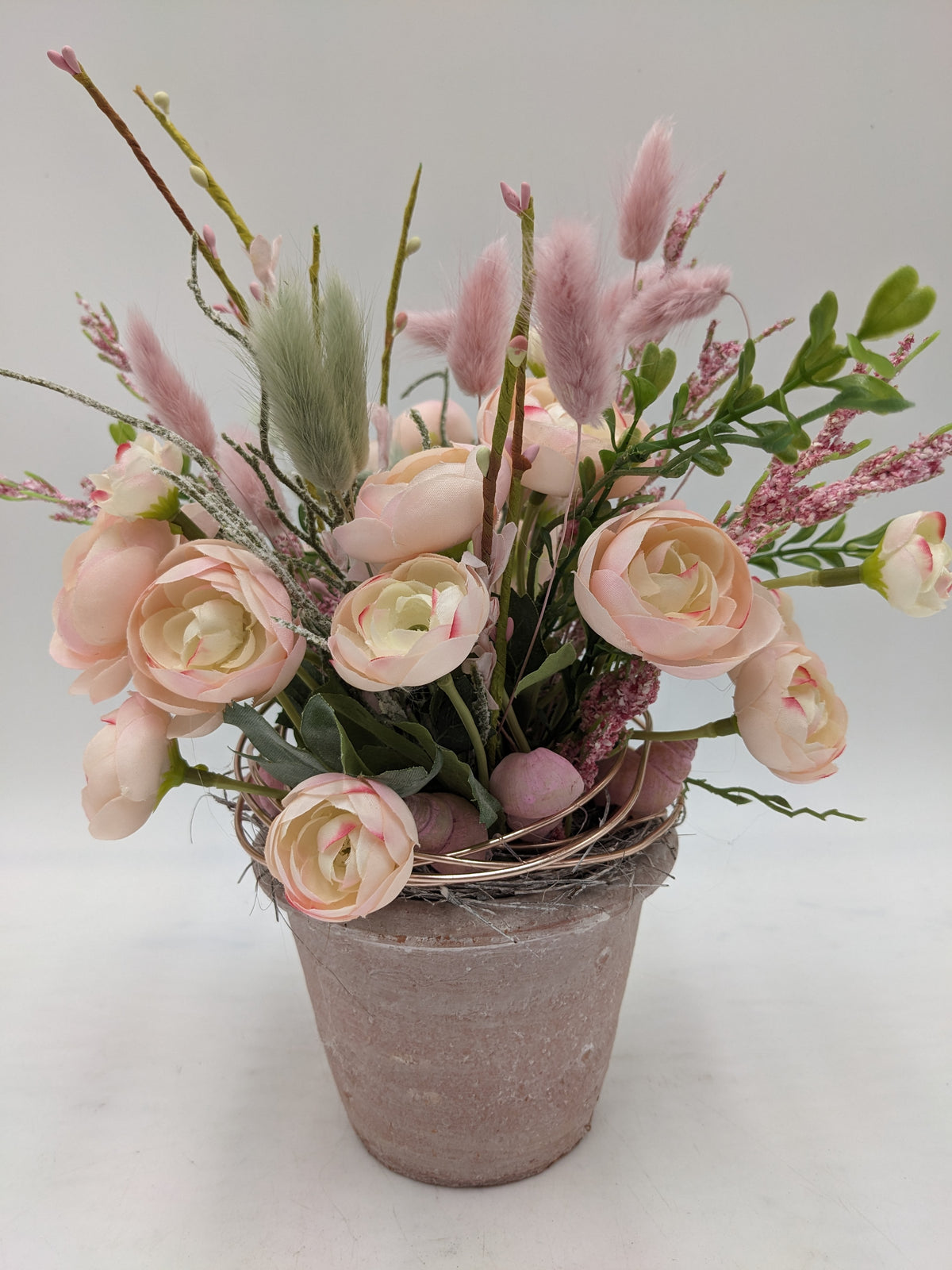 Sommergesteck mit rosa Ranunkeln, Hasenschwanzgras und Zweigen in grau-rosa Tontopf vor hellem Hintergrund