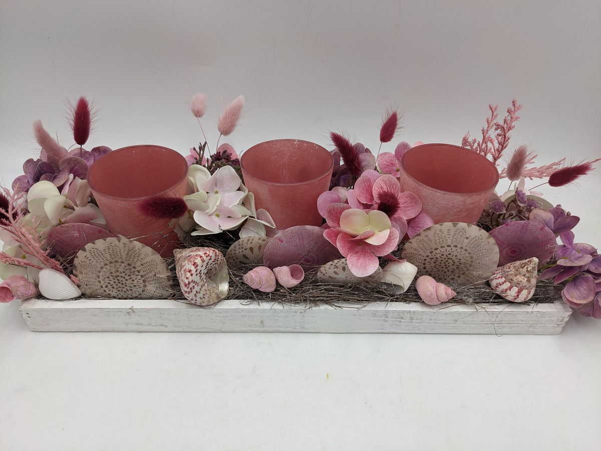 Tischgesteck in Rosa und Lila von vorn: drei rosafarbene Teelichtgläser zwischen Hortensienblüten, Schnecken und Muscheln