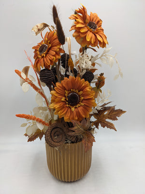 Herbstgesteck mit drei orangefarbenen Gerbera, braunen Zapfen und hellen Blättern im geriffelten Keramiktopf