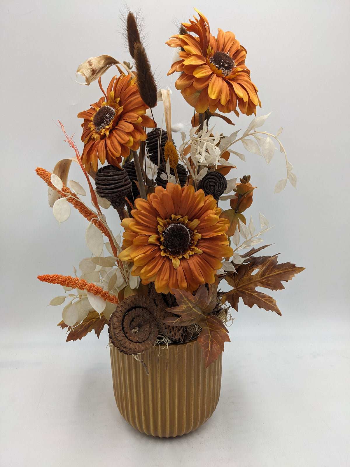 Herbstgesteck mit drei orangefarbenen Gerbera, braunen Zapfen und hellen Blättern im geriffelten Keramiktopf