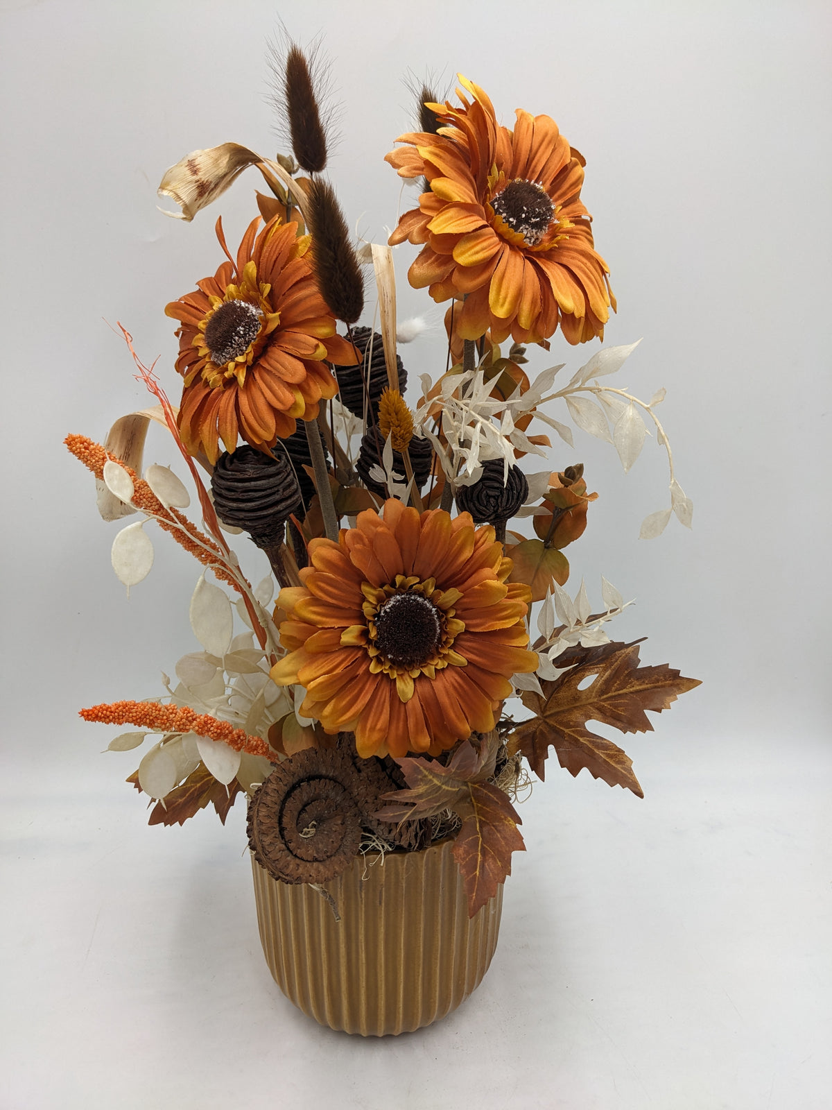 Herbstgesteck von vorn: orange Gerbera, Kolbenhirse, Gräser und braune Herbstblätter am ockerfarbenen Topf