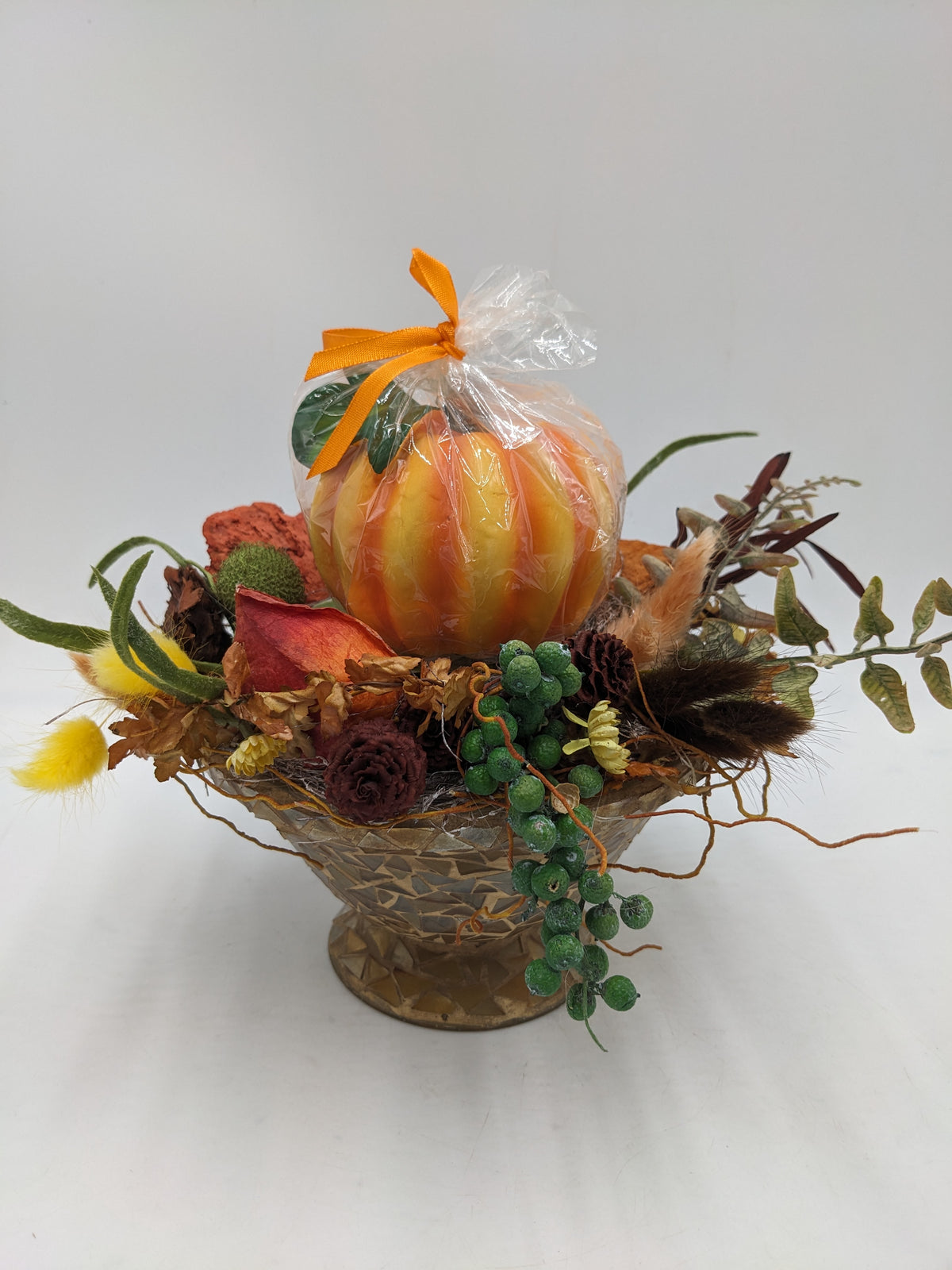 Herbstgesteck mit oranger Kürbiskerze, Zapfen, grünen Beeren und Gräsern in goldfarbener Mosaikschale