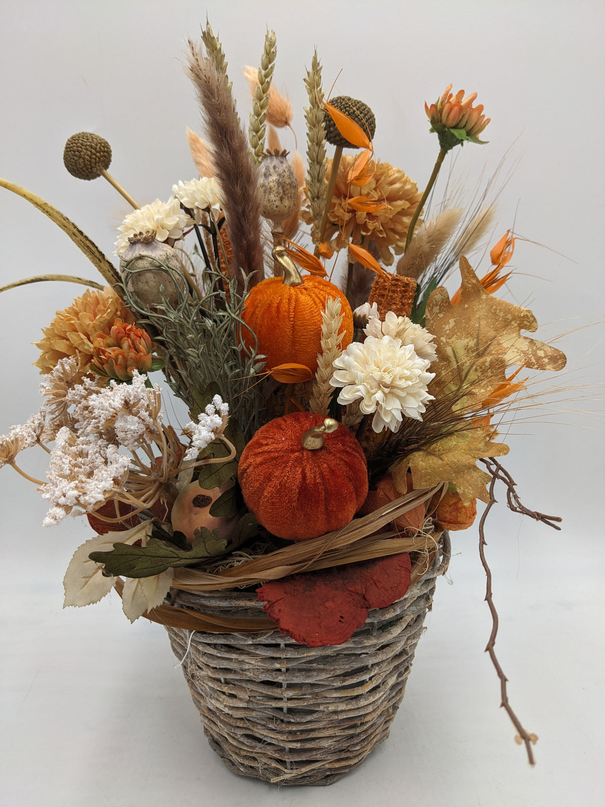Herbstgesteck im Weidenkorb mit orangefarbenen Samtkürbissen, cremeweißen Chrysanthemen, Ähren und Gräsern