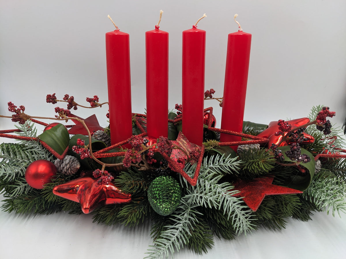 Langes Adventsgesteck mit vier roten Stabkerzen, roten Kugeln, Glassternen und Zapfen auf Tannenbeet