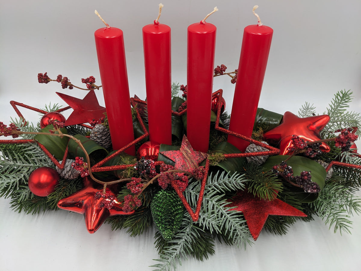 Adventsgesteck von vorn: rote Kerzen, Glitzersterne aus Draht, grüner Zapfen und rote Beeren im Tannengrün