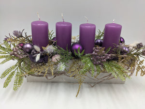 Adventsgesteck im weißen Holztablett mit vier lila Kerzen, lila Kugeln, goldenen Sternen, Beeren und Farn