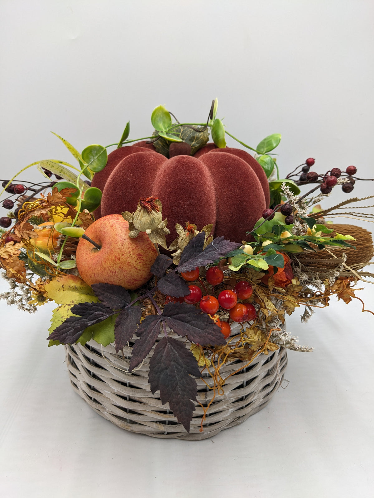 Herbstgesteck im grauen Korb mit rotbraunem Samtkürbis, Apfel, roten Beeren und dunklen Blättern