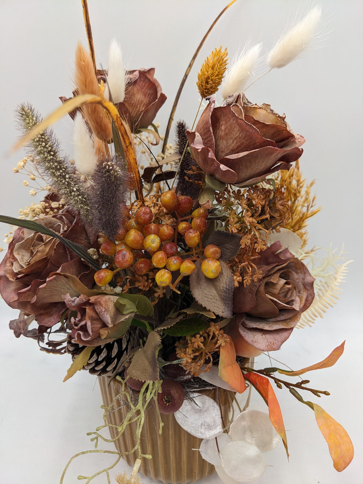 Nahaufnahme des Herbstgestecks: braune Rosen, orange-gelbe Beerendolde, helle Gräser und Silberblatt-Scheiben