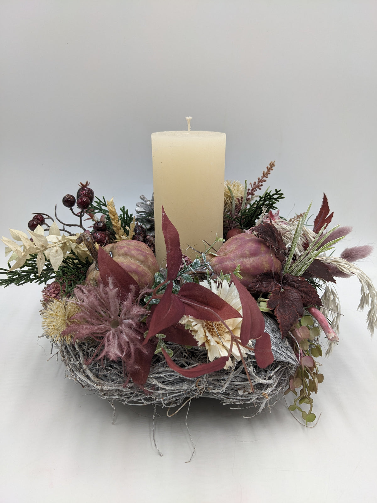 Herbstgesteck mit cremefarbener Stumpenkerze, Kürbissen, roten Beeren und dunkelroten Blättern auf hellem Geästkranz