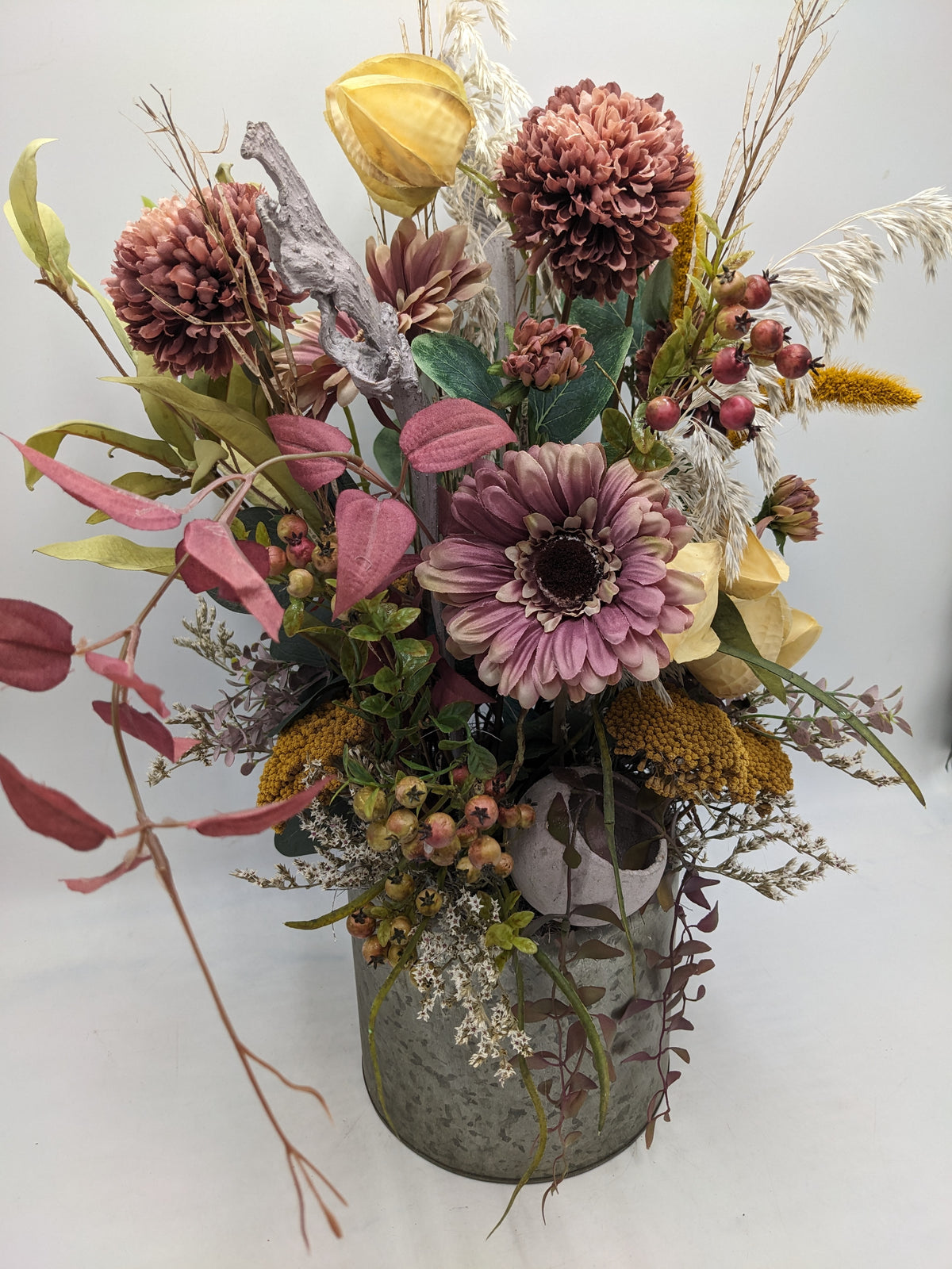 Herbstgesteck von schräg vorn: altrosa Gerbera, rote Beeren, gelbe Schafgarbe und rosa Blätter über grauem Zinktopf