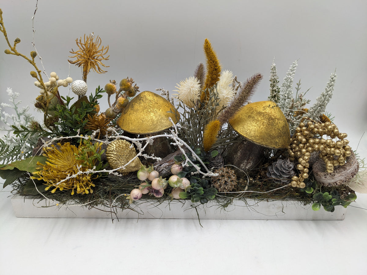 Langes Herbstgesteck im weißen Holztablett mit zwei goldenen Pilzen, goldenem Igel, Disteln und Gräsern