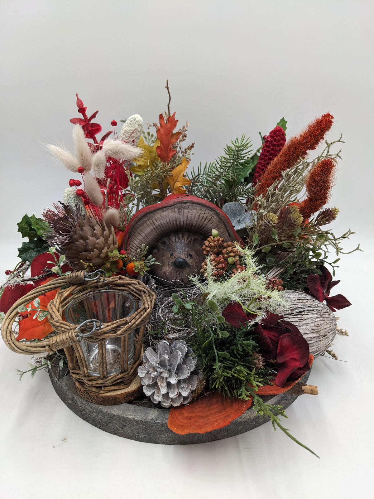 Herbstgesteck auf grauem Holztablett mit Keramik-Igel, Weidenwindlicht, Zapfen, Gräsern und roten Rispen