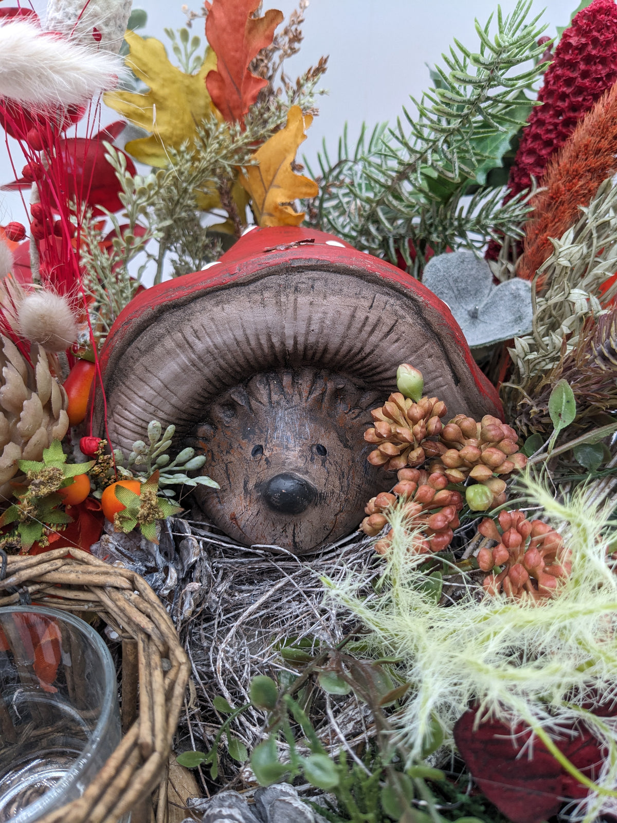 Nahaufnahme des Keramik-Igels mit roter Pilzkappe im Zweignest, umgeben von Beeren, Herbstblättern und Moos