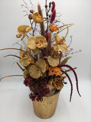 Herbstgesteck von vorn: Orchideenblüten, dunkelrote Ähren und beige Gräser in goldfarbenem Topf