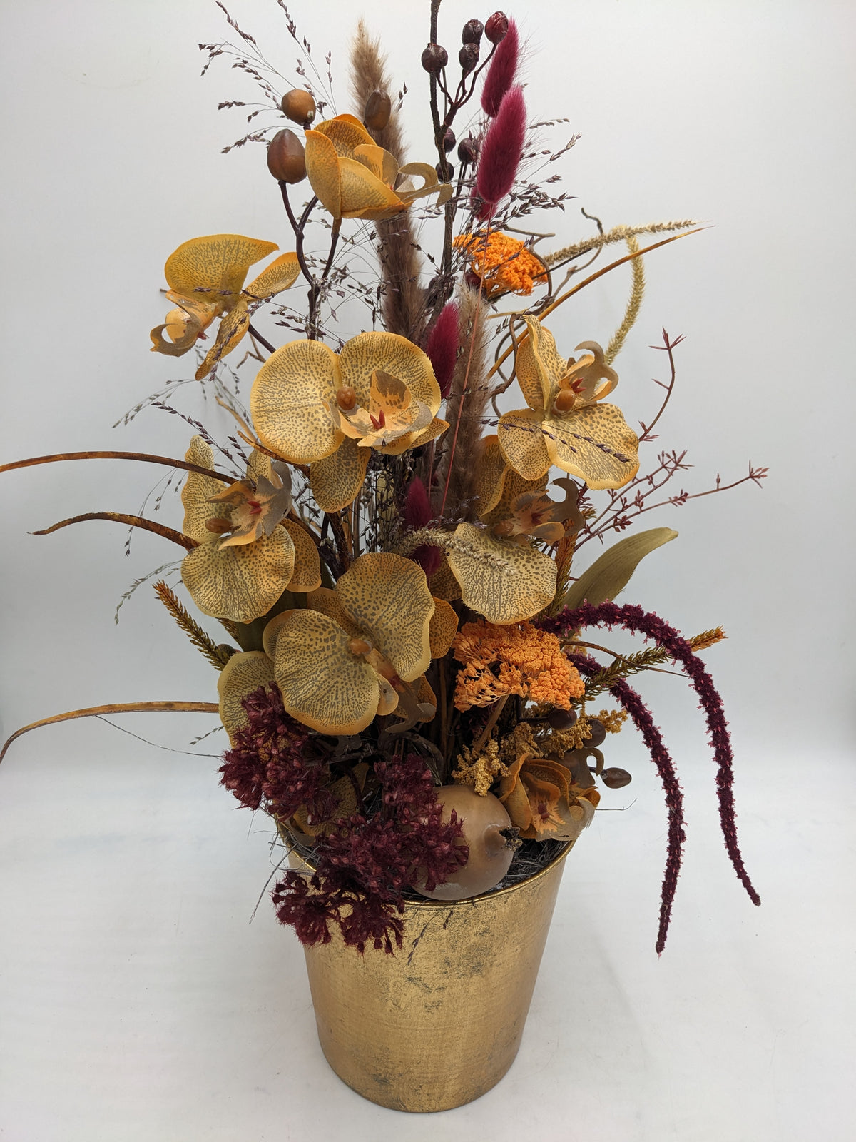 Herbstgesteck von vorn: Orchideenblüten, dunkelrote Ähren und beige Gräser in goldfarbenem Topf