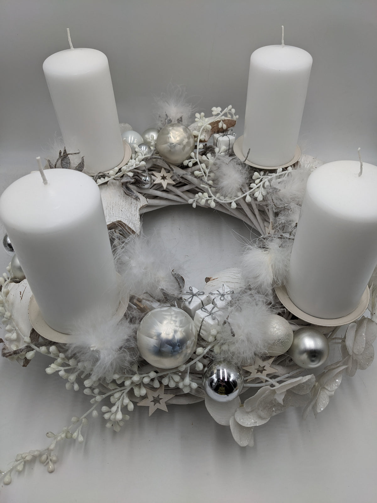 Adventskranz von oben: heller Rattankranz mit vier weißen Kerzen, silbernen Kugeln, Federn und Geschenkpäckchen