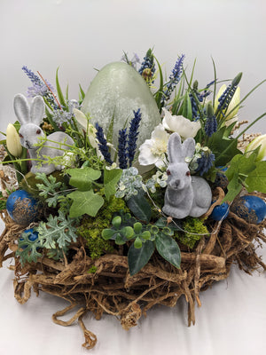 Ostergesteck mit grünem Osterei, zwei grauen Samthasen, blauen Hyazinthen und weißen Blüten in Wurzelschale
